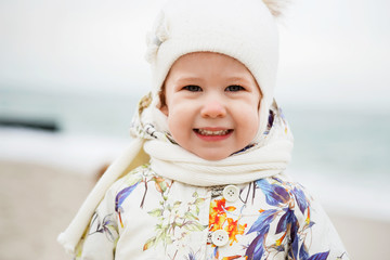 Cute little girl playing on the sandy beach. Happy child wearing warm floral print jacket, pom pom hat and scarf playing outdoors on fall, winter or spring day.