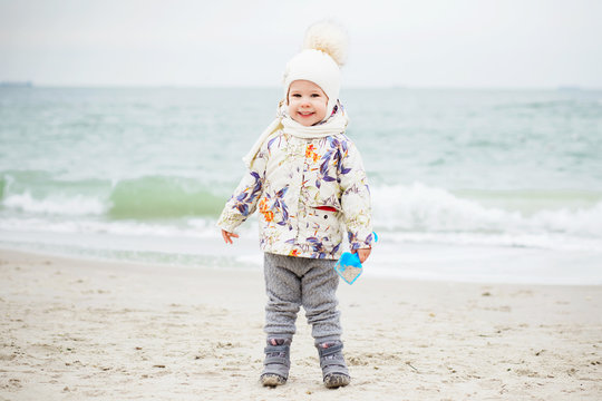 Cute little girl playing on the sandy beach. Happy child wearing warm floral print jacket, pom pom hat and scarf playing outdoors on fall, winter or spring day.