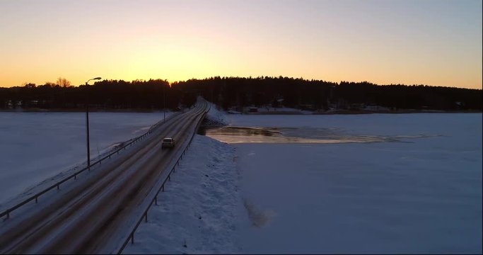 Car on a winter bridge, Cinema 4k aerial view following car on a frozen archipelago bridge, revealing the sunset above a town, on a sunny winter evening dawn, in sarkisalo, Varsinais-suomi, Finland