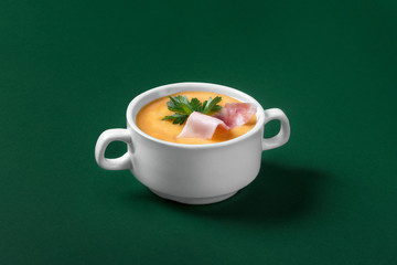 Cream soup is yellow with bacon and parsley on a green background. Side view. Deep white plate with handles