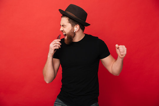 Image of joyous guy wearing black t-shirt and hat singing and listening to music with cell phone and wireless earphones, isolated over red wall