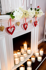 A cozy white fireplace with lots of candles in glass with hanging red hearts on it.