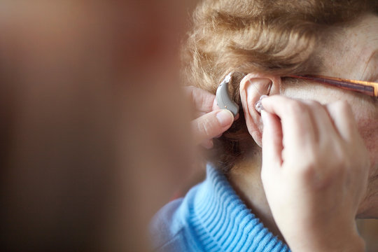Mature woman helping senior woman insert hearing aid, close-up, differential focus