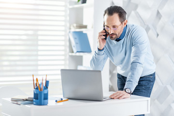 Obraz premium Pleasant talk. Handsome alert dark-haired bearded man talking on his phone and looking at the screen of his laptop while standing near the table