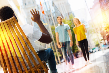 Unexpected meeting. Pleasant young couple having a relaxed walk while noticing their friend sitting in a little cafe and waving to them