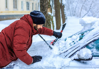 Young man clean car after snow storm with scraper - obrazy, fototapety, plakaty