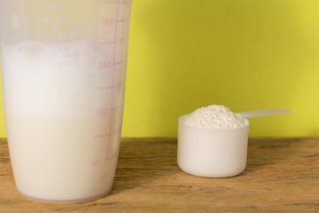 Whey Protein. Front view of white scoop with vanilla flavour powder and shaker on rustic wooden table. Solid color: yellow.