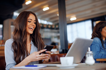 Young woman texting on the phone with a laptop on table in cafe