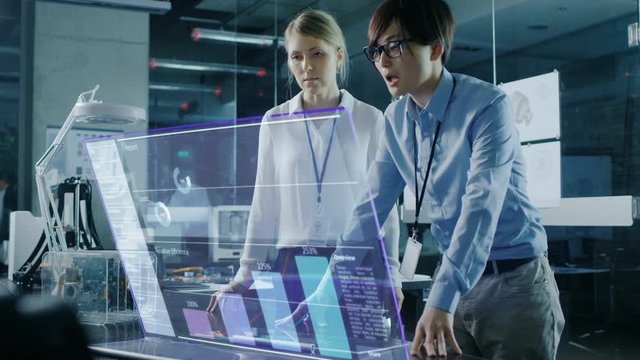 Male and Female Business People Have Discussion while Using Modern Computer With Transparent Holographic Display. Monitor Shows User Interface with Interactive Charts and Graphics.