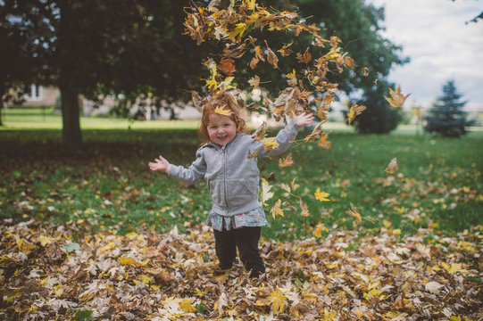 Portrait of red haired female toddler in park throwing autumn leaves