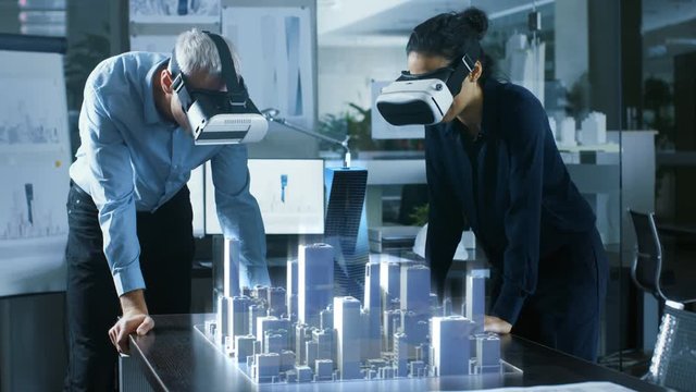 Male and Female Architects Wearing  Augmented Reality Headsets Work with 3D City Model. High Tech Office Professional People Use Virtual Reality Modeling Software Application.