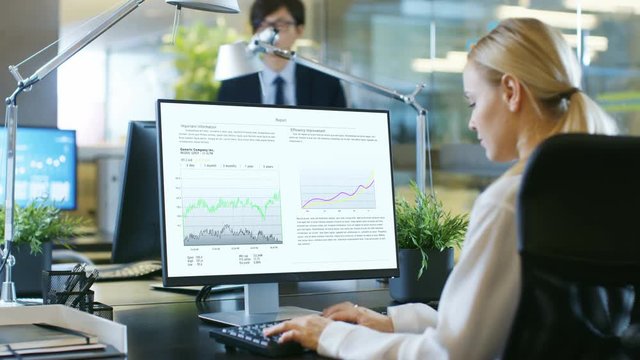 In the Office Businesswoman Works on Personal Computer with Spreadsheets, Charts and Statistics. Her Colleague sits Opposite to Her. Room is Stylishy made of Concrete and Glass.