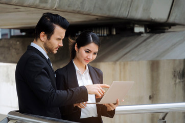 two business asian woman and caucasian man in suit talking and reading information about finance news in laptop computer in modern city, network technology, internet, successful and teamwork concept