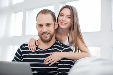 Fototapeta premium My beloved. Attractive glad long-haired young woman hugging her handsome boyfriend and smiling while he using a laptop