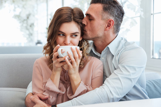 Picture of adult caring man hugging and kissing his girlfriend in city cafe, while woman drinking cappuccino from cup