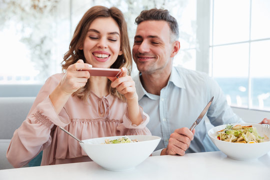 Photo of cheerful married couple having dinner, and woman photographing her meal on smartphone before eating