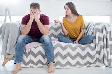 Cheating on her. Attractive heart-broken young woman talking to her man about his unfaithfulness and he closing his face while sitting on the couch - obrazy, fototapety, plakaty