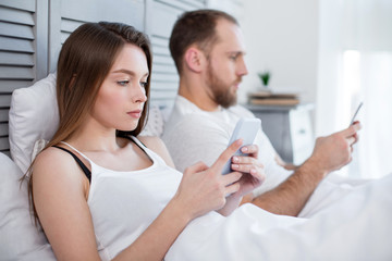 Ready for bed. Beautiful concentrated unsmiling dark-haired woman typing on her phone while lying in bed and her man using his phone while lying next to her