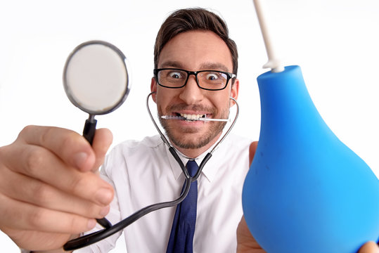 Handsome bearded scarry doctor with  syringe in his mouth with stethoscope in his ears and a douchebag in his hand isolated on a white background is smiling wearing glasses