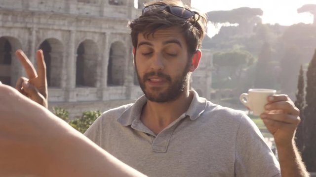 Beautiful attractive young man with blue eyes and brown beard posing for funny pictures with cup of coffee in front of colosseum in rome at sunset