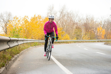 Fototapeta na wymiar Young Woman in Pink Jacket Riding Road Bicycle on the Highway in the Cold Autumn Day. Healthy Lifestyle.