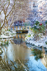 Fototapeta na wymiar Small river with mini pont in the park, outdoor in Paris, France, during snowfall in winter. Taken in the morning with sunrise light. Smart concept of modern park for family recreation.