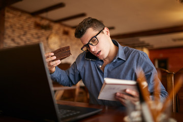 Fototapeta na wymiar Overworked businessman speaking on cellphone holding it with shoulder, reading writings in notebook, eating a cake and working on laptop simultaneously. Multitasking concept. Busy stylish man.