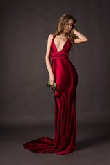 Beautiful blonde girl wearing a deep neckline red satin dress holding red rose in her hand on a grey background