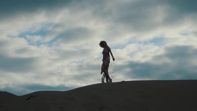Silhoutte of a fabulous woman standing and posing on sand hill outdoor. Wind is blowing on her half naked body.