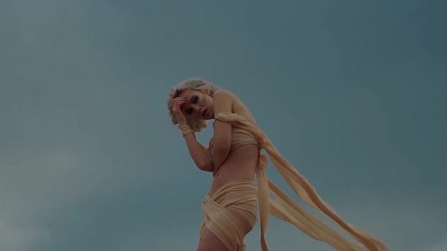 Shot from below of a beautiful girl posing in a silk pieces of cloth on a cloudy day. Silk stripes are floating on a wind.