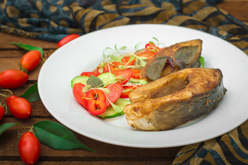Grilled fish steak with vegetables on plate: tomatoes, microgran, cucumber, tasty and healthy dinner. Wooden rustic background. Top view