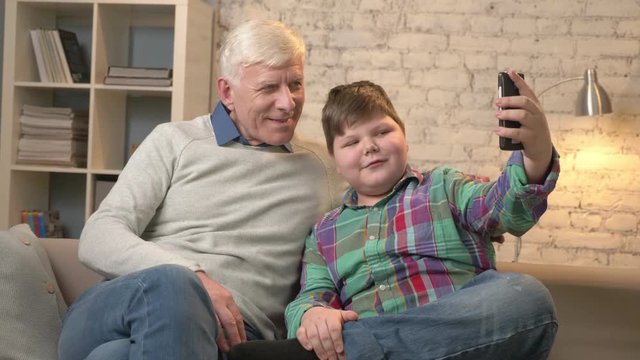 Grandfather and grandson are sitting on the sofa using a smartphone, do selfie. Young fat child and grandfather. boy shows the sign of peace. Home comfort, family idyll, cosiness concept. 60 fps