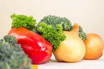 Different vegetables and fresh herbs with waterdrops on the white table. Parsley and green broccoli sprouts, raw not-pealed onion, red sweet pepper in the center