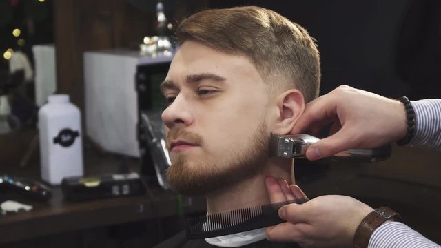 Close up of a handsome young man smiling while professional barber trimming his beard with a clipper trimmer equipment service consumerism masculinity beardcut barbering barbershop lifestyle.