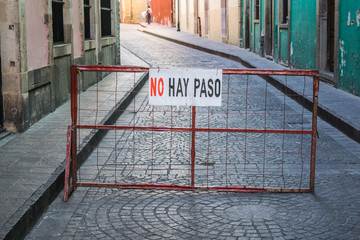 A red iron gate with a no passing sign, blocking the entrance to a cobblestone street, with the bottom portion of colorful buildings, in the background, in Guanajuato, Mexico - 192048969