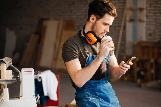 Carpenter Man using smart phone and holding cup of coffee in front of table with tools at workshop