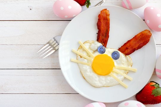Easter breakfast with cute bunny face made of egg and bacon. Flay lay scene with decor over a white wood background.