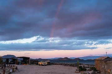 A rainbow over a homestead on the Baja California Peninsula. The sea of cortez is in background. An old, yellow truck at sunrise with cactus and garage
