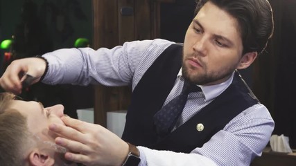 Professional barber working at his barbershop shaving his young male client with a razor careful dangerous professionalism experience skills barbering styling beard.