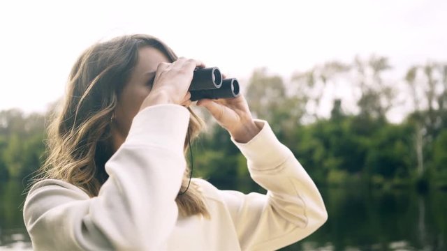 Beautiful young woman in a white cardigan is using a pair of binoculars while riding on a boat in a river. Handheld real time medium shot