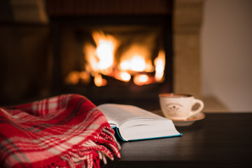 Wool warm blanket or plaid and a book with a cup of tea on the background of burning fireplace