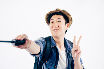 Young Asian tourist with hat smiling and holding selfie stick taking a selfie photo isolated over...