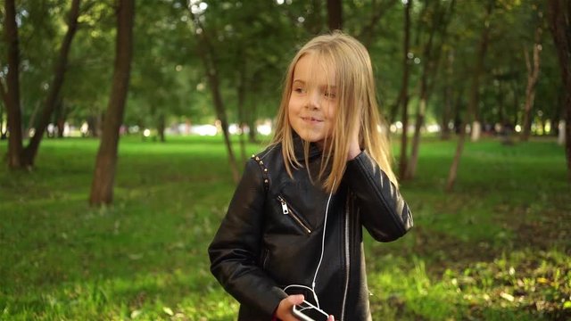 Cute little blonde girl wearing a leather jacket listening to the music standing in a park on a summer day. Locked down real time medium shot