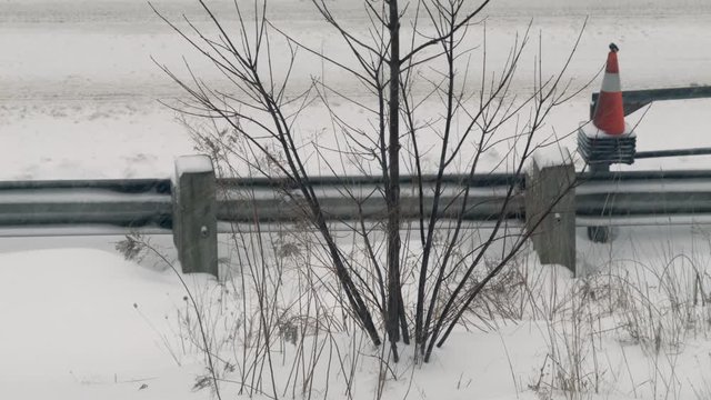 Heavy snow falling along guardrail of highway with orange road pylons.