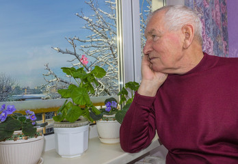 Old lonely man sitting near the window in his house.