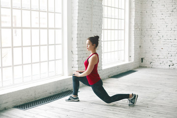 Confident determined student girl with hair knot doing physical workout indoors before university. Stylish sporty young woman in sneakers and sportswear standing in low lunge, stretching leg muscles