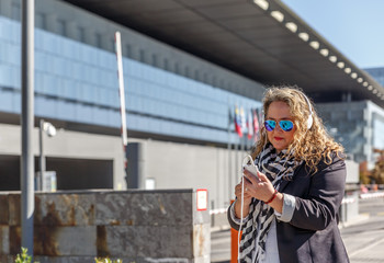 A blonde woman with black glasses and casual clothes listens to music with her headphones and checks her mobile phone