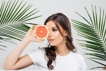 Portrait of a smiling woman with brunette hair posing with green leaves and holding sliced grapefruit isolated over white background