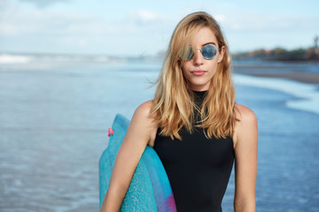 Good looking blonde young woman wears trendy shades and swimsuit, holds surfboard, ready to conquer ocean waves, poses against nice sea lanscape, blue sky. Surfer lifestyle and water activities