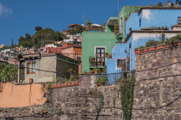 A hill with a stone wall at the bottom and colorful houses climbing up the hill, some greenery and a clear blue sky, in Guanajuato, Mexcio - 192074327